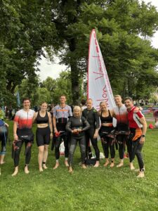 Triahtlon Väst, Ironman Jönköping 2026 Åtta triathleter från Triahtlon Väst står på rad på en gräsmatta i en park, barfota och med våtdräkten nerdragen till midjan. Bakom dem står en vit och röd vimpel med texten "sim.cykel.löp - sedan -89". I bakgrunden syns tävlingsområde för Ironman Jönköping och lövträd, mulet väder.