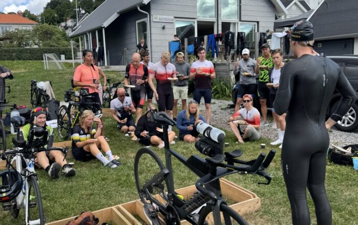 Pontus, i våtdräkt, visar växlingsteknik för en grupp triathleter som sitter i gräset. Framför dem står en tempocykel i ett bäxlingsområde med cykelställ, med skor och utrustning utlagda.