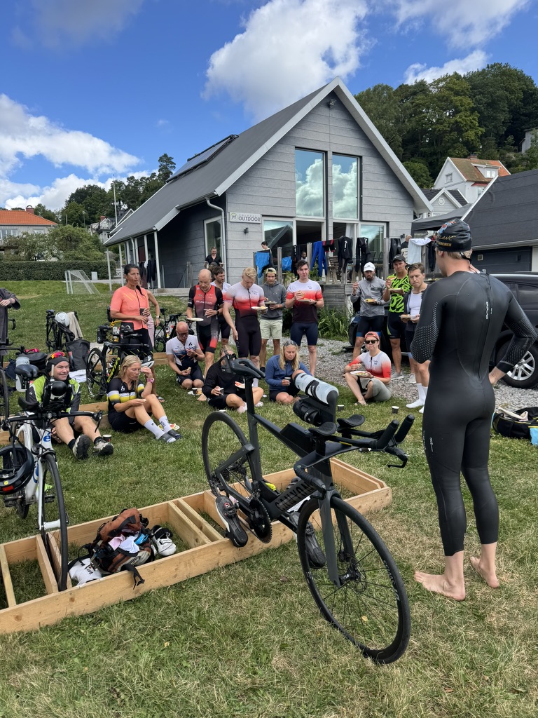 Pontus, i våtdräkt, visar växlingsteknik för en grupp triathleter som sitter i gräset. Framför dem står en tempocykel i ett bäxlingsområde med cykelställ, med skor och utrustning utlagda.